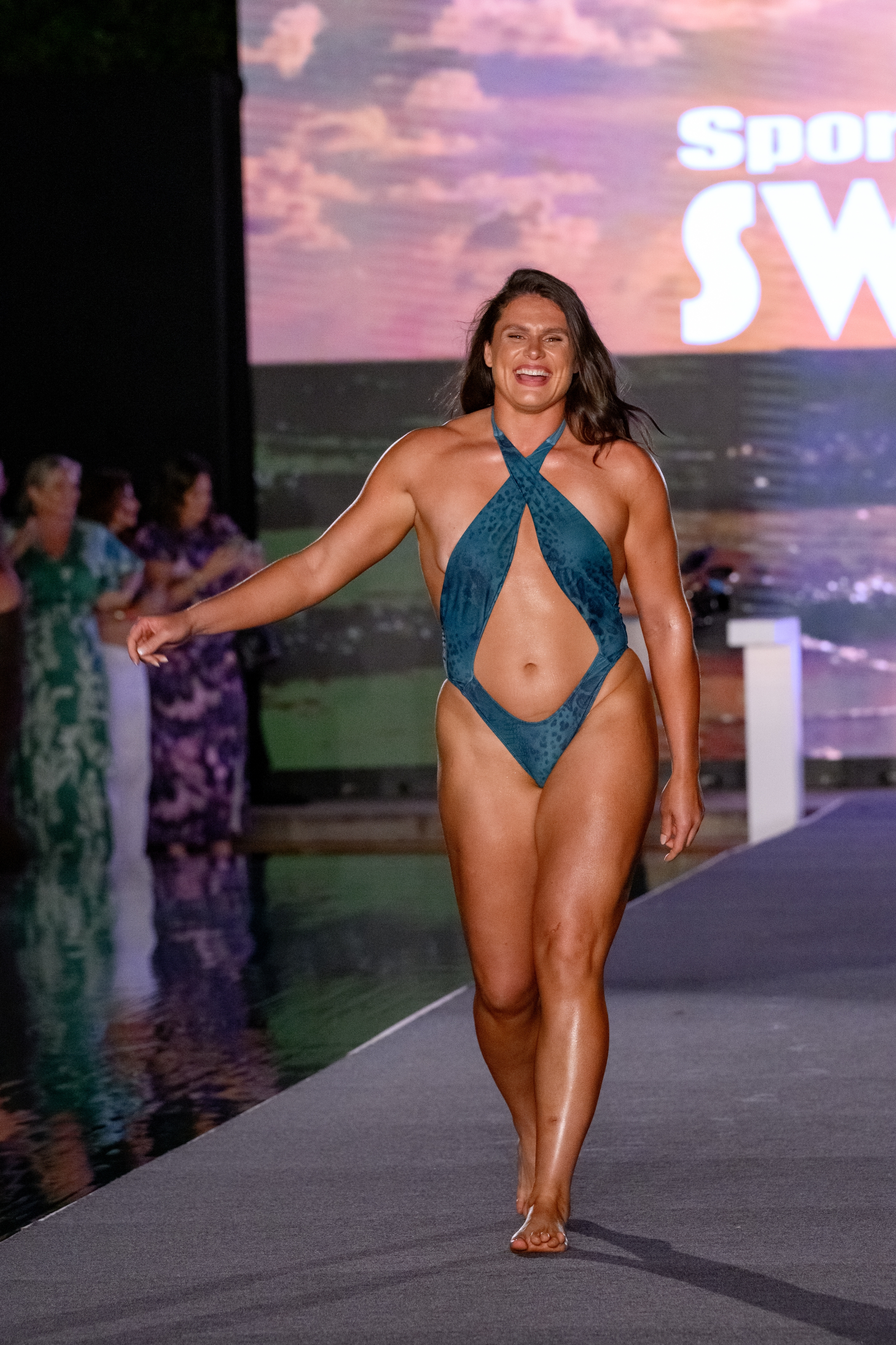 Person modeling a stylish, cut-out, one-piece swimsuit on a runway. Background features a "Sports" sign. Others in similar attire are visible