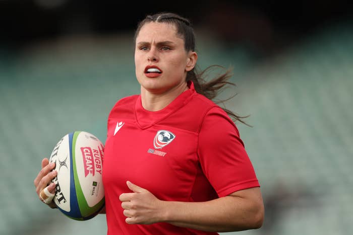 Person wearing a sports jersey holds a rugby ball on a field, looking towards the camera with determination