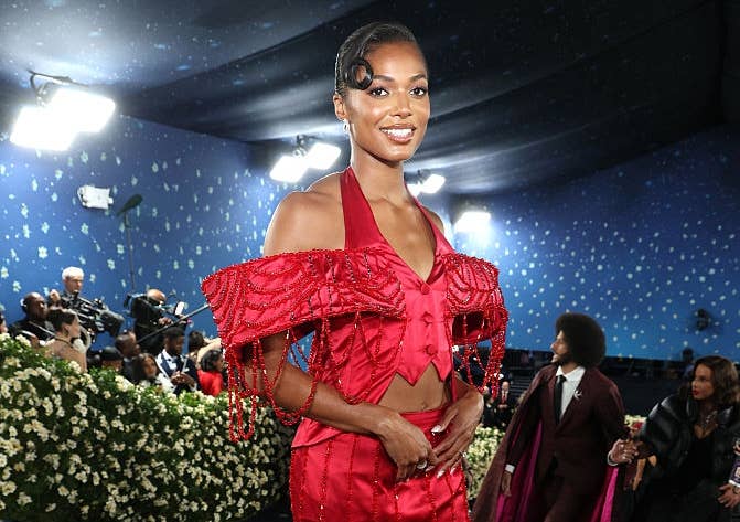 A person in an elegant, off-shoulder red gown with intricate details, posing on the red carpet surrounded by photographers