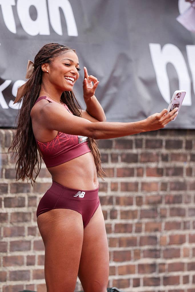 Person in athletic wear takes a selfie, smiling and gesturing with one hand, in front of a brick wall