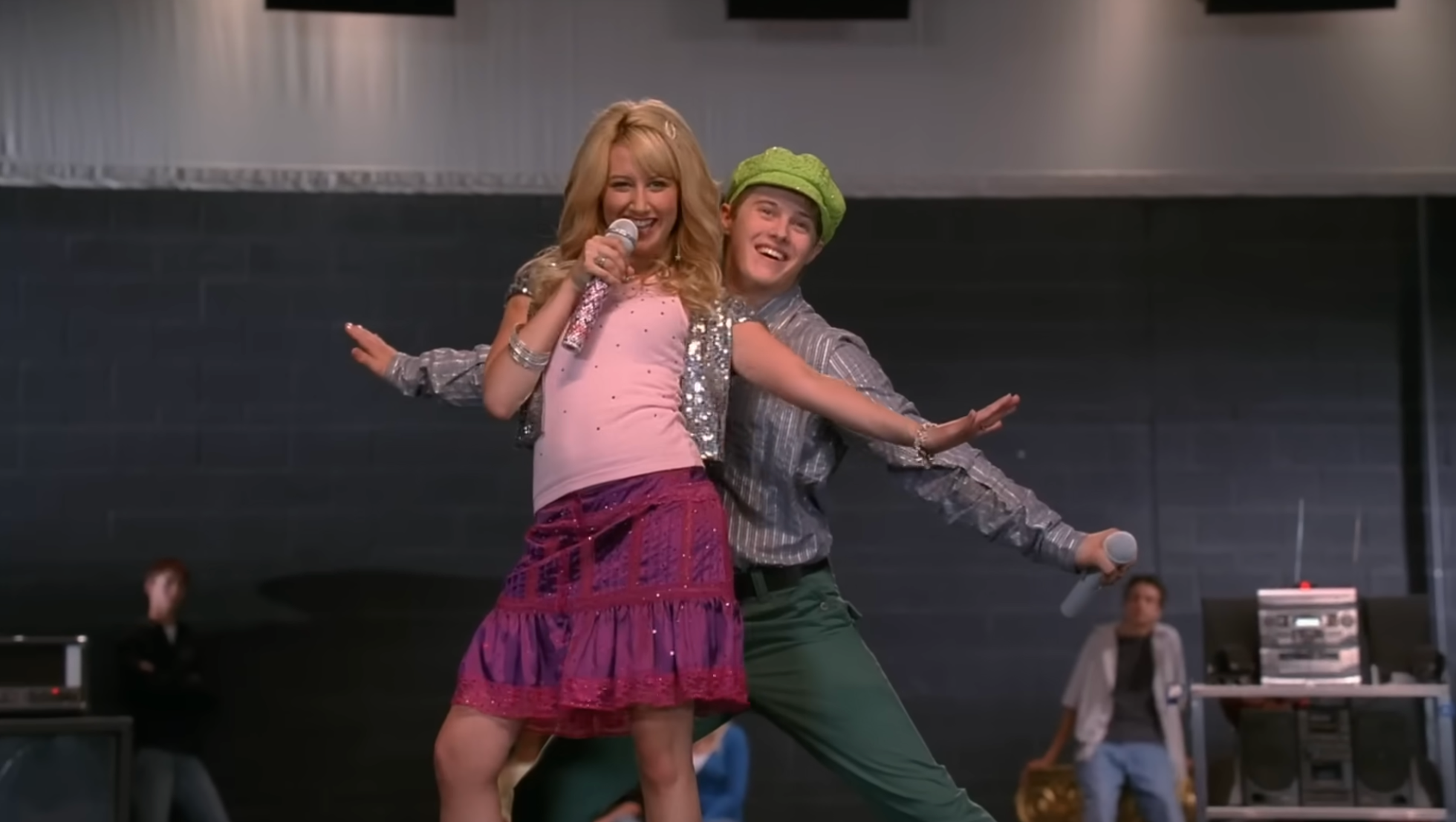 Two people perform energetically on stage, one with a microphone and sparkly outfit, and the other in a hat with a striped shirt and green pants