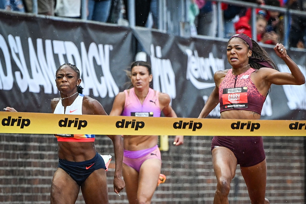 Athletes cross the finish line in an intense track race, showing determination. They wear athletic gear as the crowd watches in the background