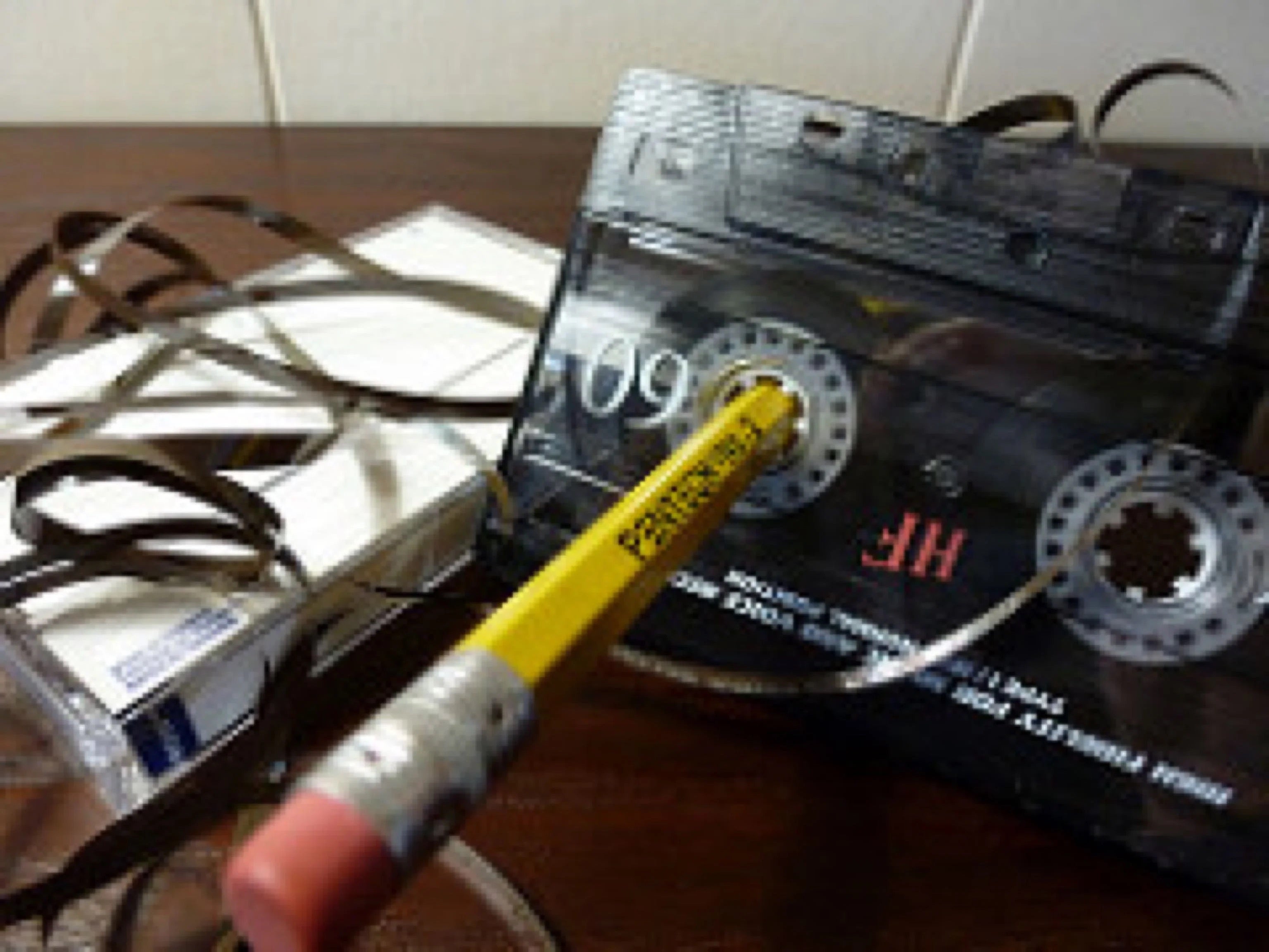 Cassette tape with unwound tape strands, a pencil inserted in one reel, and empty cassette cases in the background