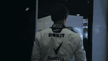 A person in a racing suit walks down a corridor, displaying team logos like &quot;ROKiT&quot; and &quot;Rexona&quot; on the back