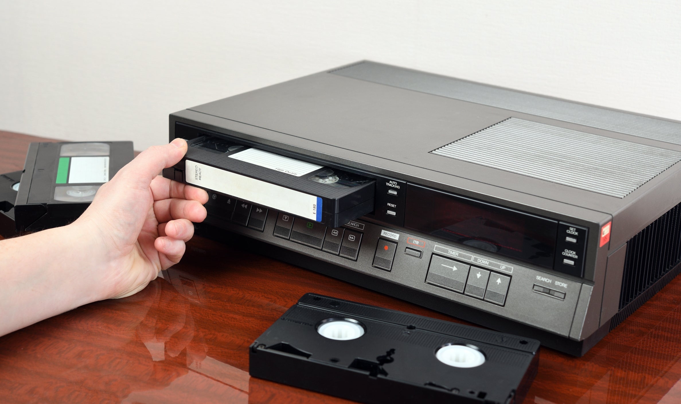 Person inserting VHS tape into a VCR, with additional tapes on a wooden surface