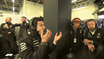 People in a Formula 1 pit garage wearing team jackets and headsets, engaged in animated discussion. Equipments and screens surround them