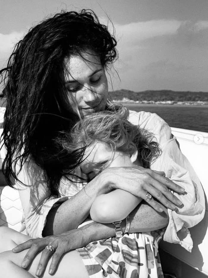 Woman embraces child on a boat, sharing a tender moment. Woman has long hair and wears a casual shirt; the child is in a patterned dress