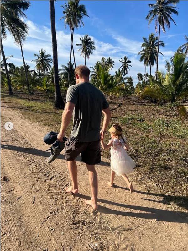 Man and young girl walk barefoot on a sandy path, surrounded by palm trees. He carries a helmet and shoes, and she wears a white dress