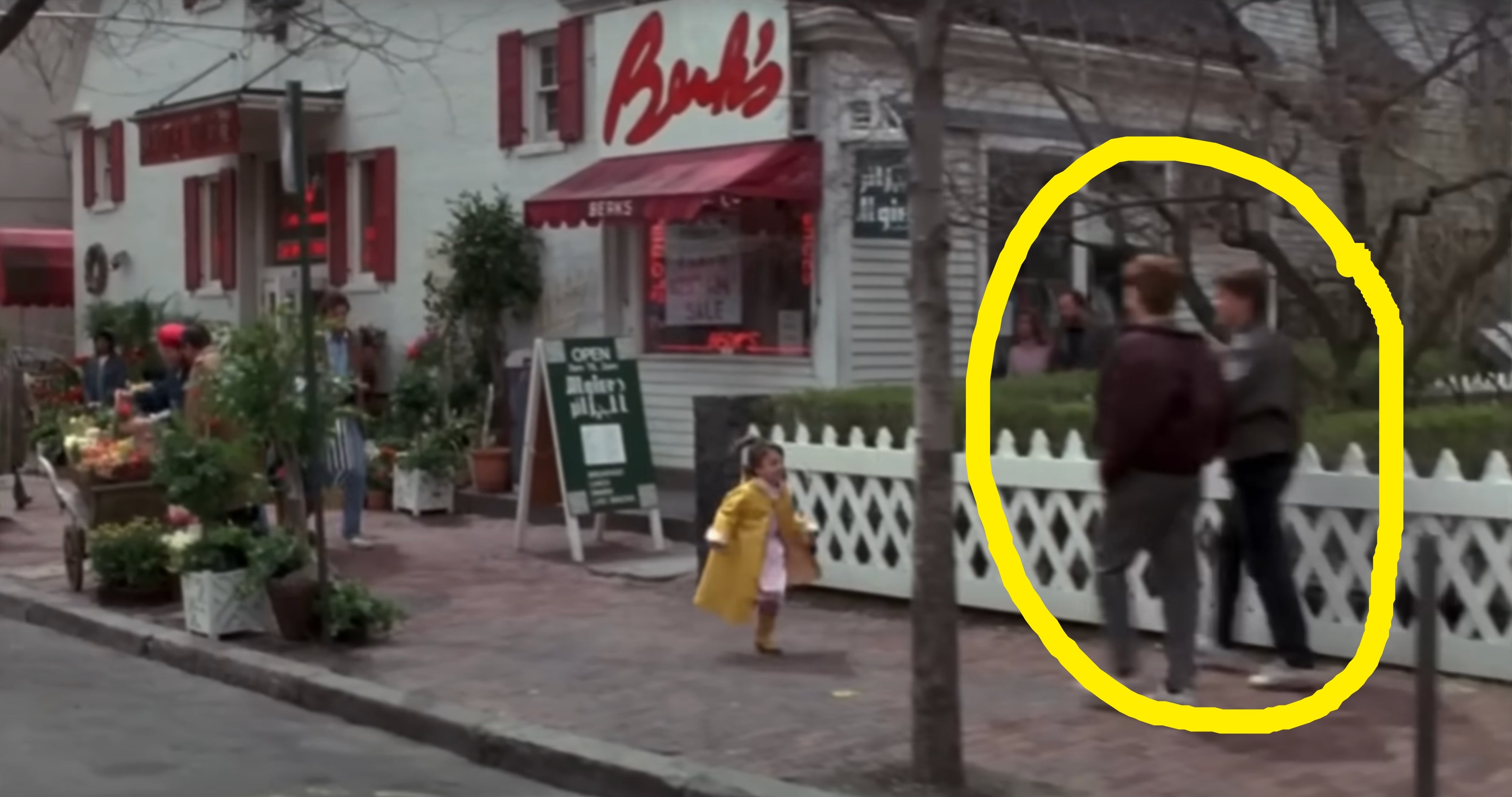 Child in yellow outfit walks towards adults on sidewalk near a market with fresh produce and a store with "Brab's" sign in a small-town setting