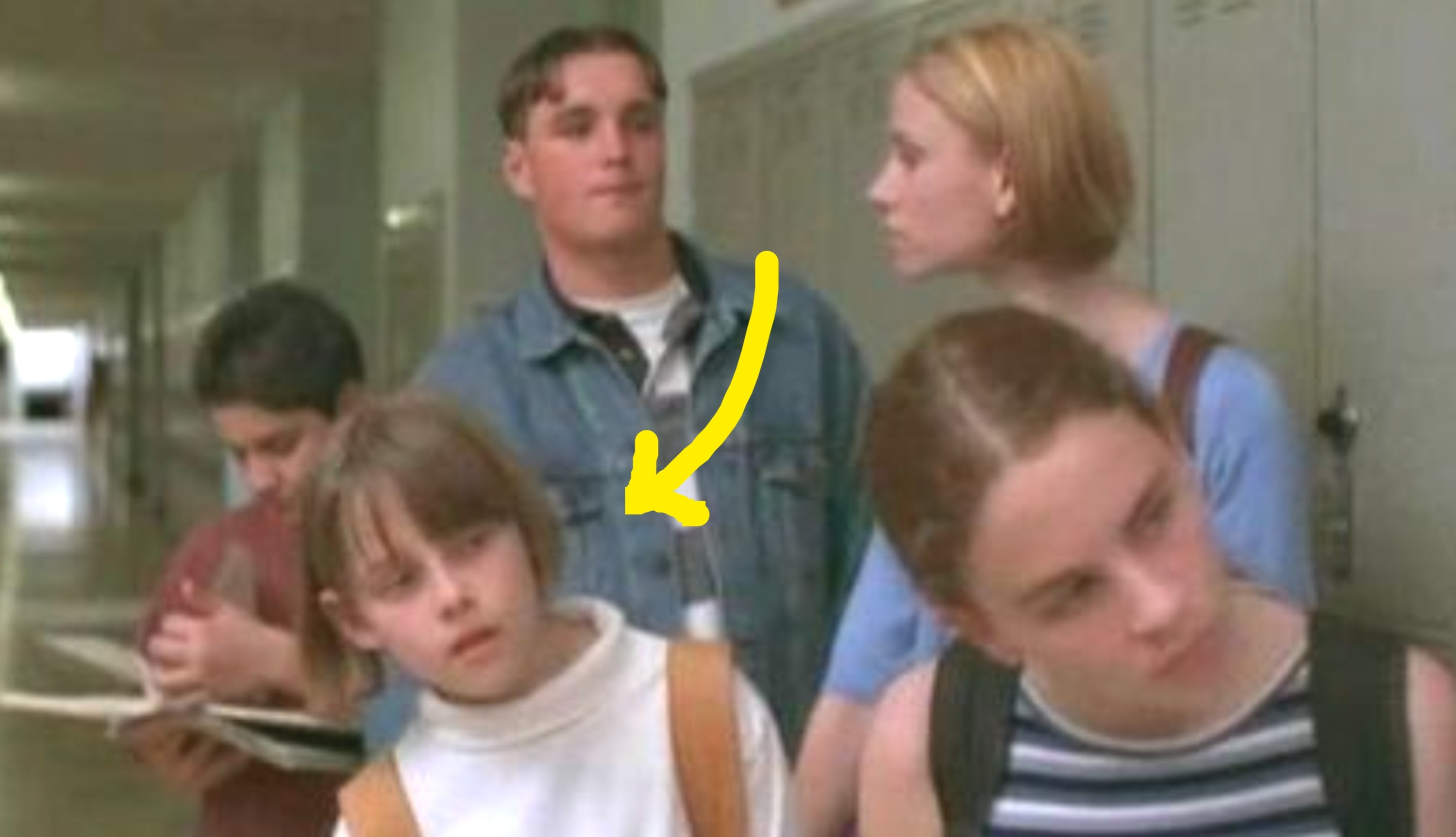 Group of teens in a school hallway, casually dressed, some carrying backpacks. One is holding a book, appearing to chat and navigate lockers