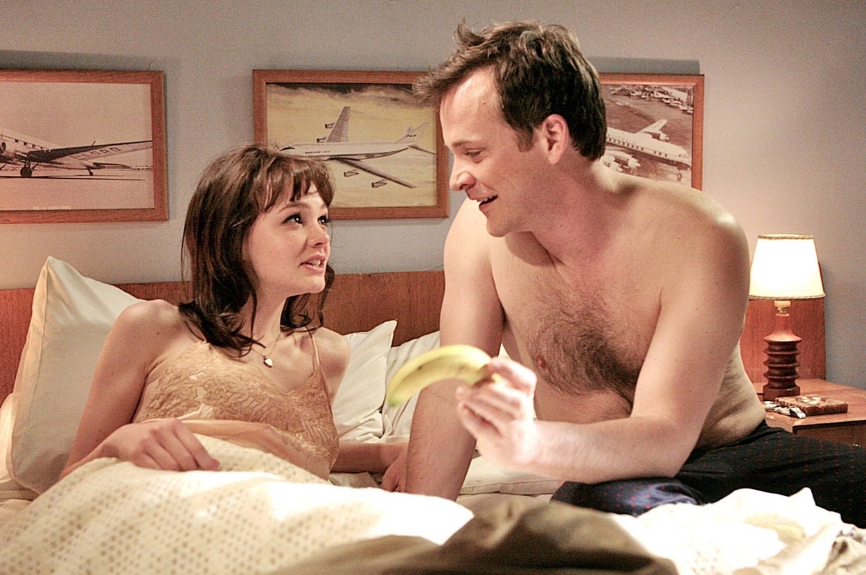A couple in bed shares a lighthearted moment; the man humorously holding a banana, both seated under airplane wall art