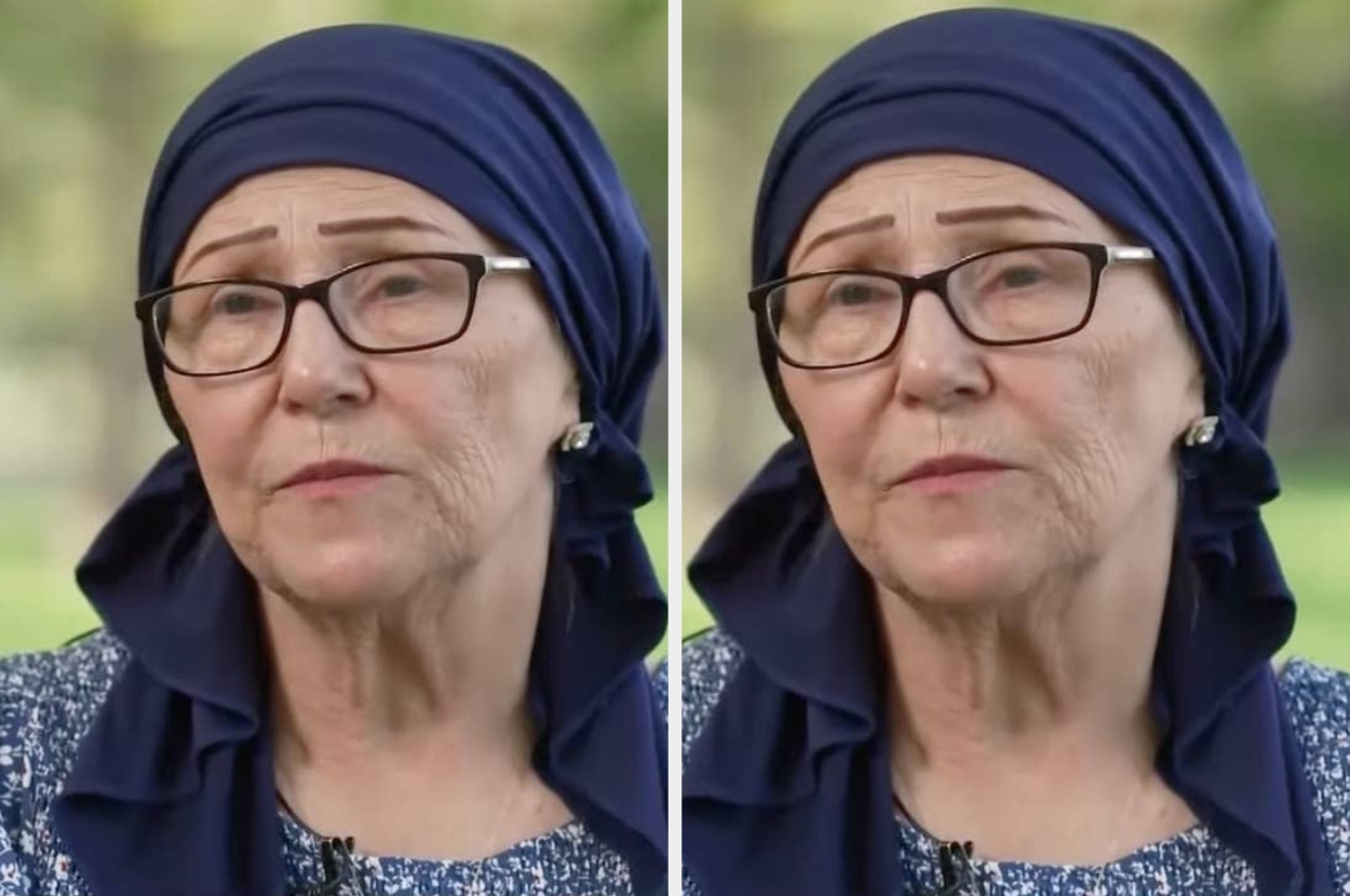 An older woman wearing glasses and a headscarf speaks in an outdoor setting, looking thoughtful