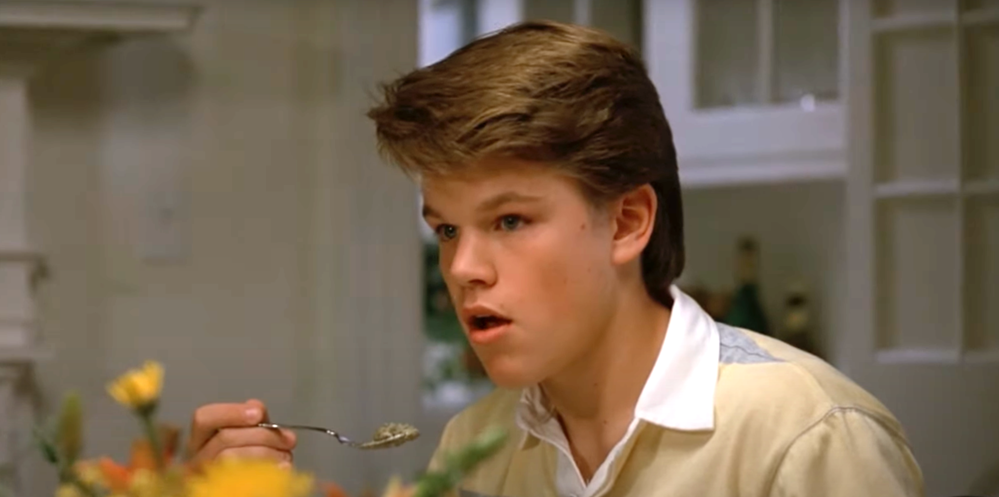 Young individual with short hair, surprised expression, holds a spoon near mouth while seated at a dining table indoors