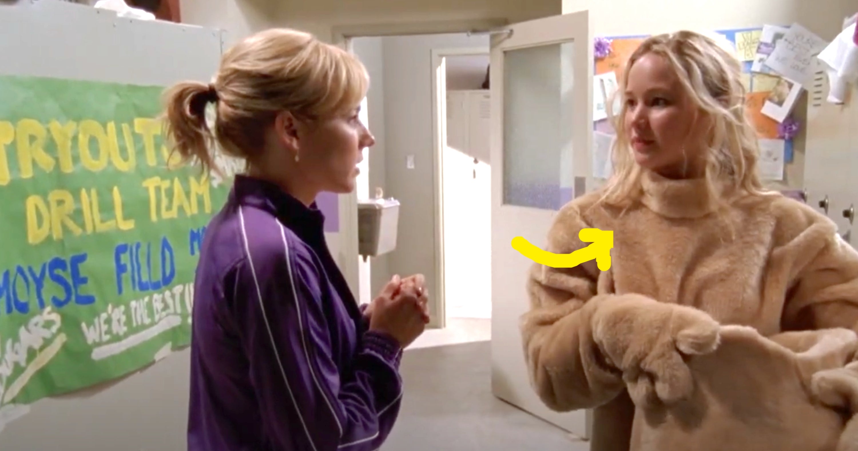 Two women talk in a hallway. One wears a purple tracksuit; the other is in a fuzzy, oversized outfit. A tryouts poster hangs on the wall