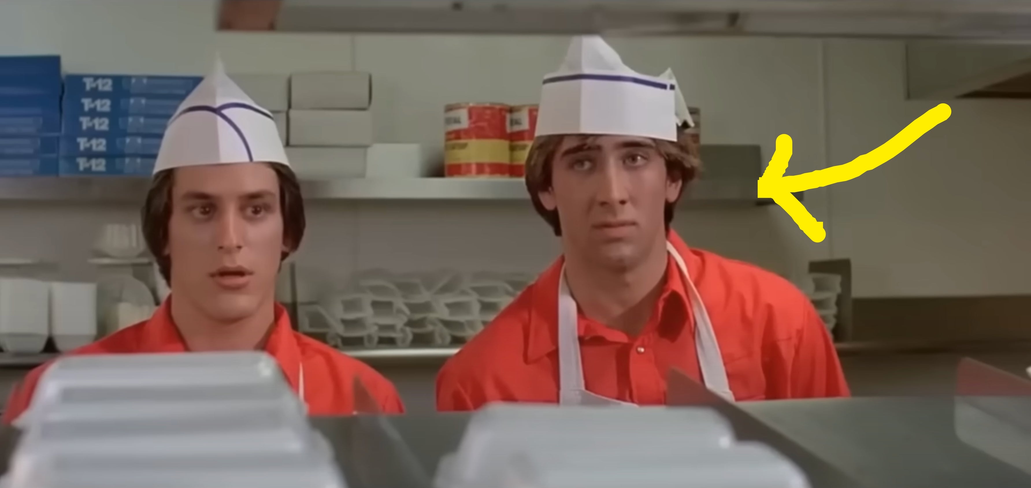 Two people in a fast-food kitchen wearing red uniforms and paper hats, looking surprised