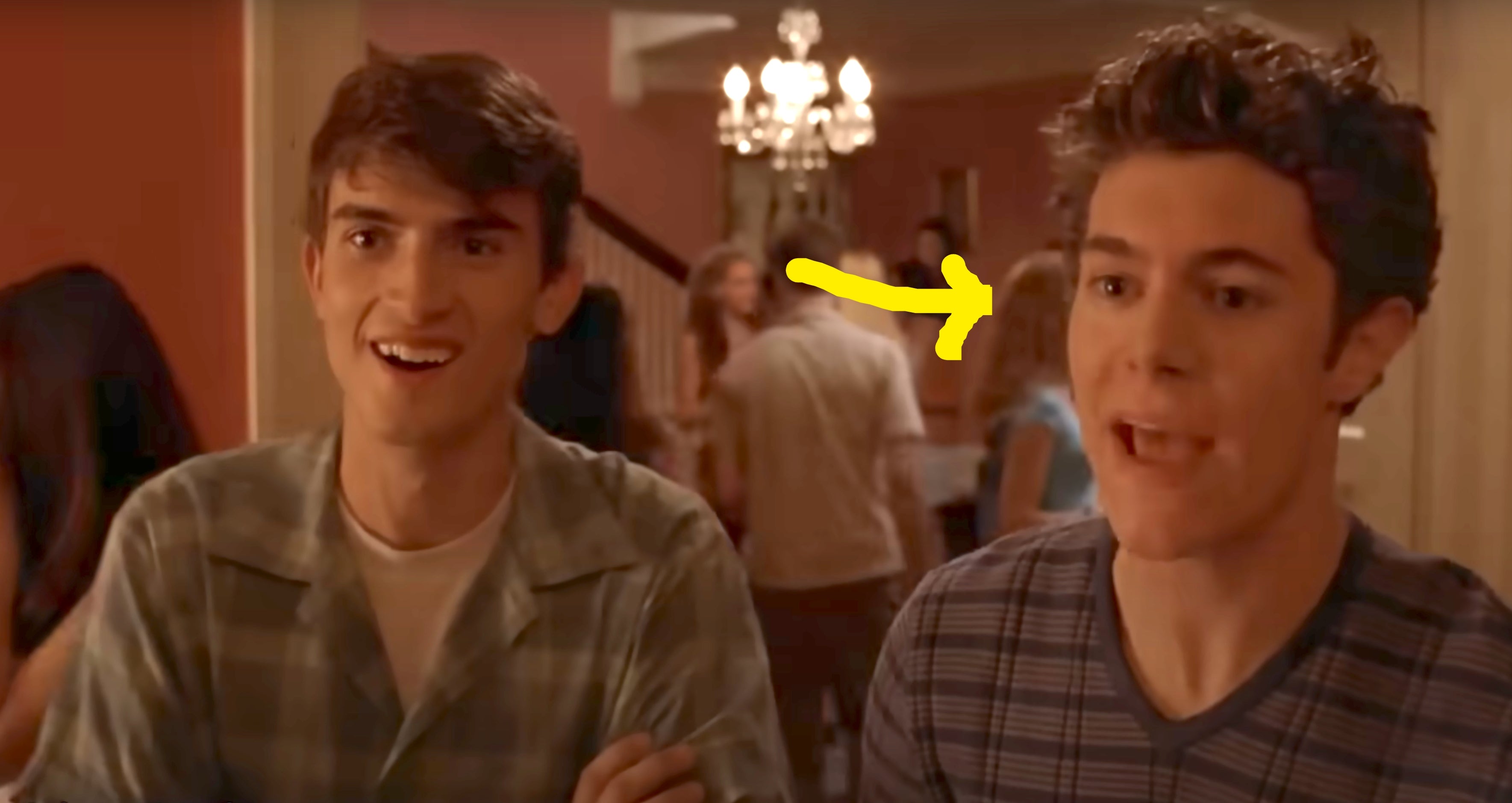 Two young men are laughing at a party indoors. One wears a plaid shirt, and the other a striped shirt. A chandelier is visible in the background