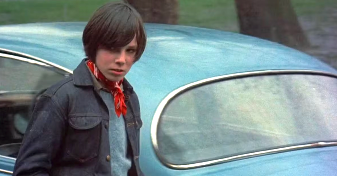 Person with short hair stands beside a vintage car, wearing a denim jacket and red scarf