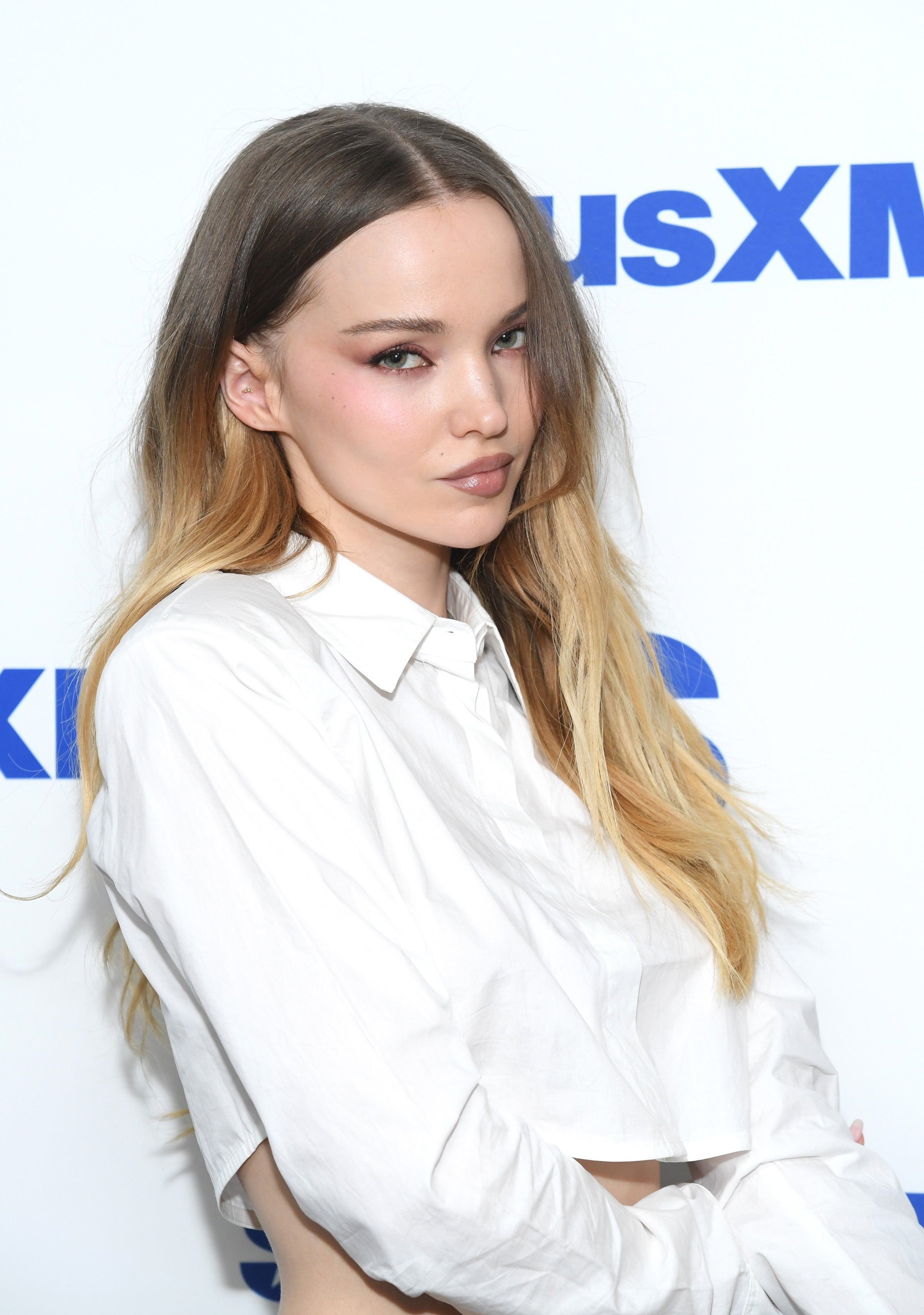 Person with long hair wearing a cropped white button-up shirt poses against a SiriusXM backdrop