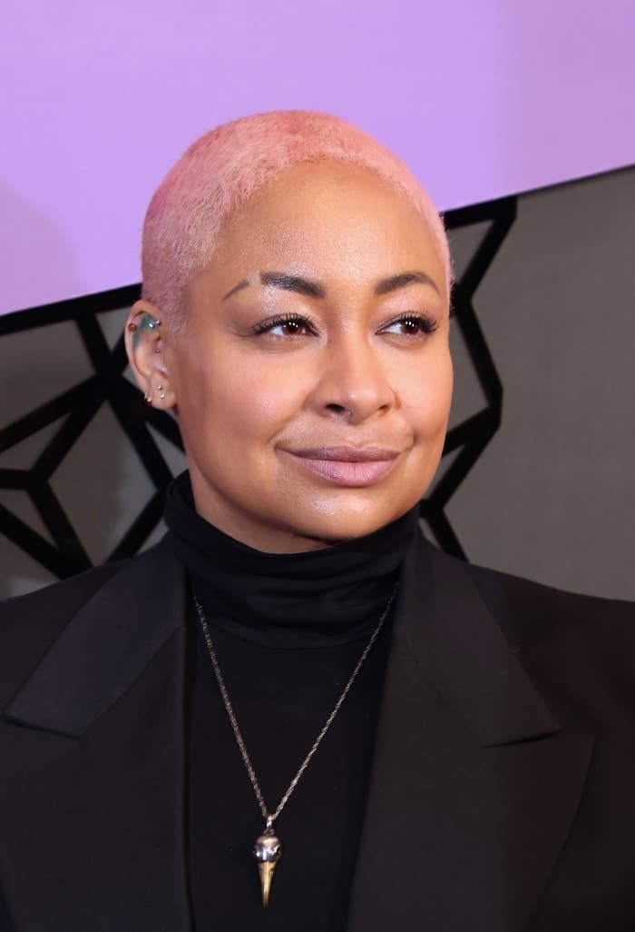 Person with short hair wearing a blazer and turtleneck poses confidently against a geometric wall