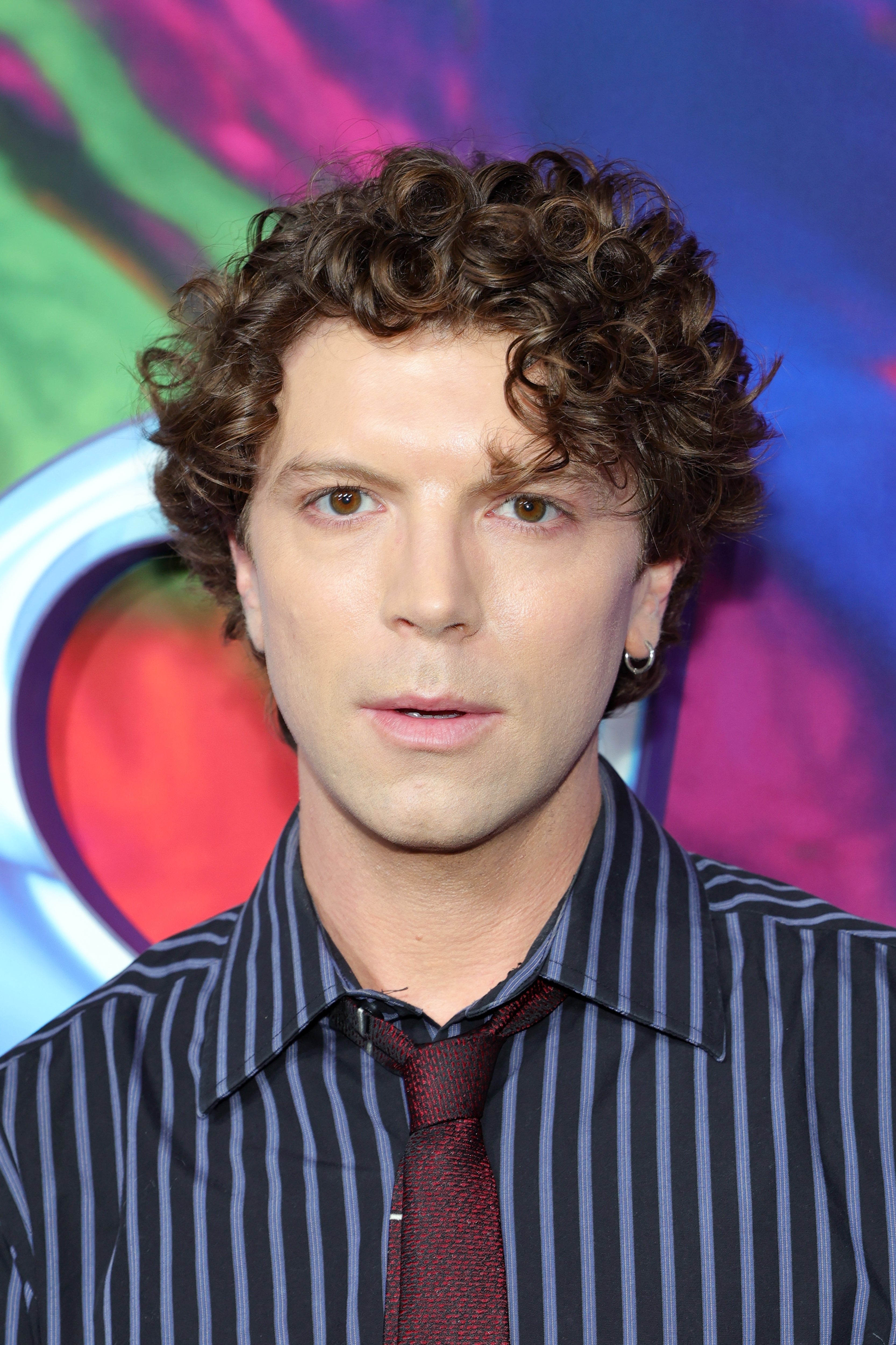 Person with curly hair wears a striped shirt and tie, posing at an event with a vibrant background