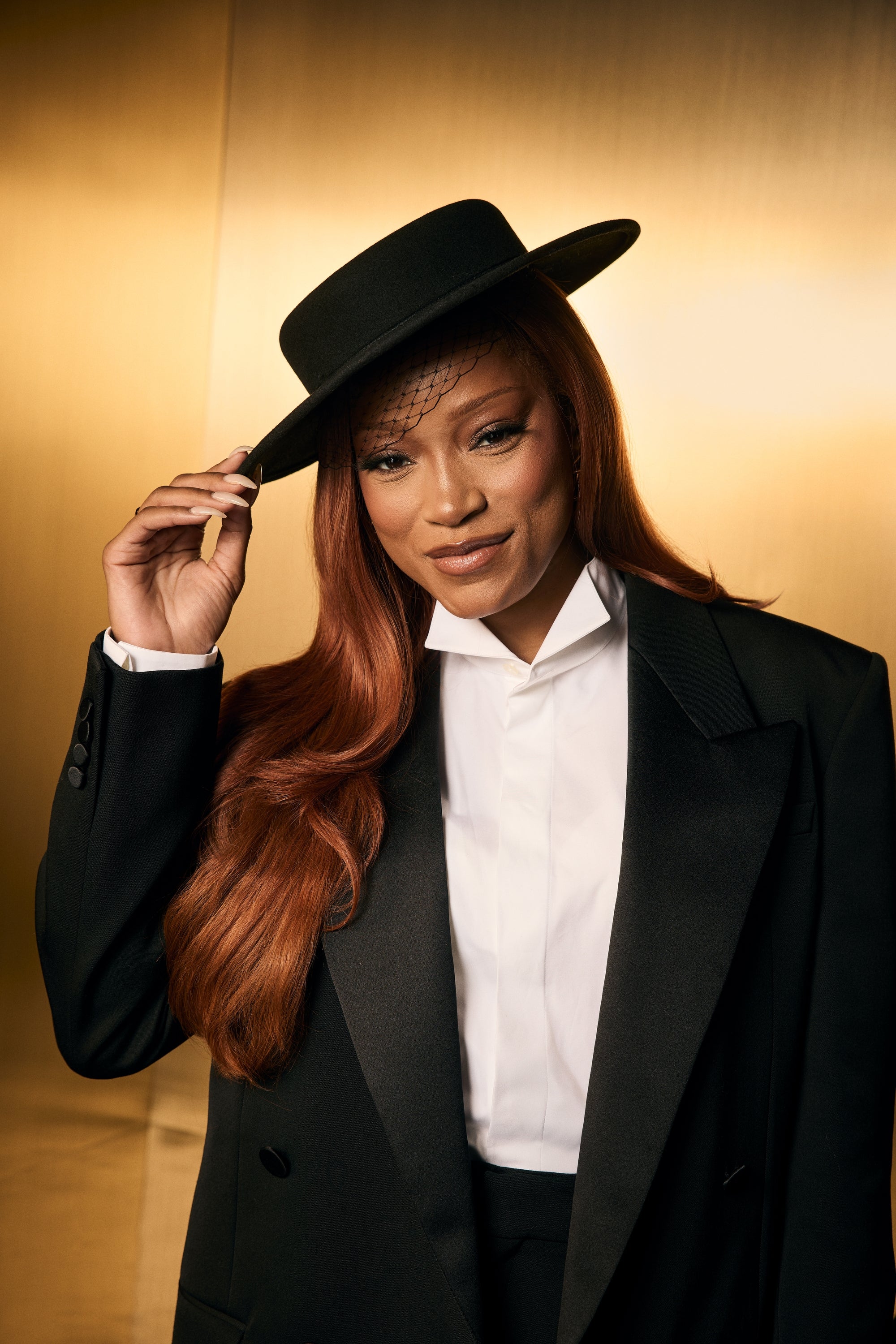 Person wearing a tailored suit and hat with a netted veil, posing confidently against a simple backdrop
