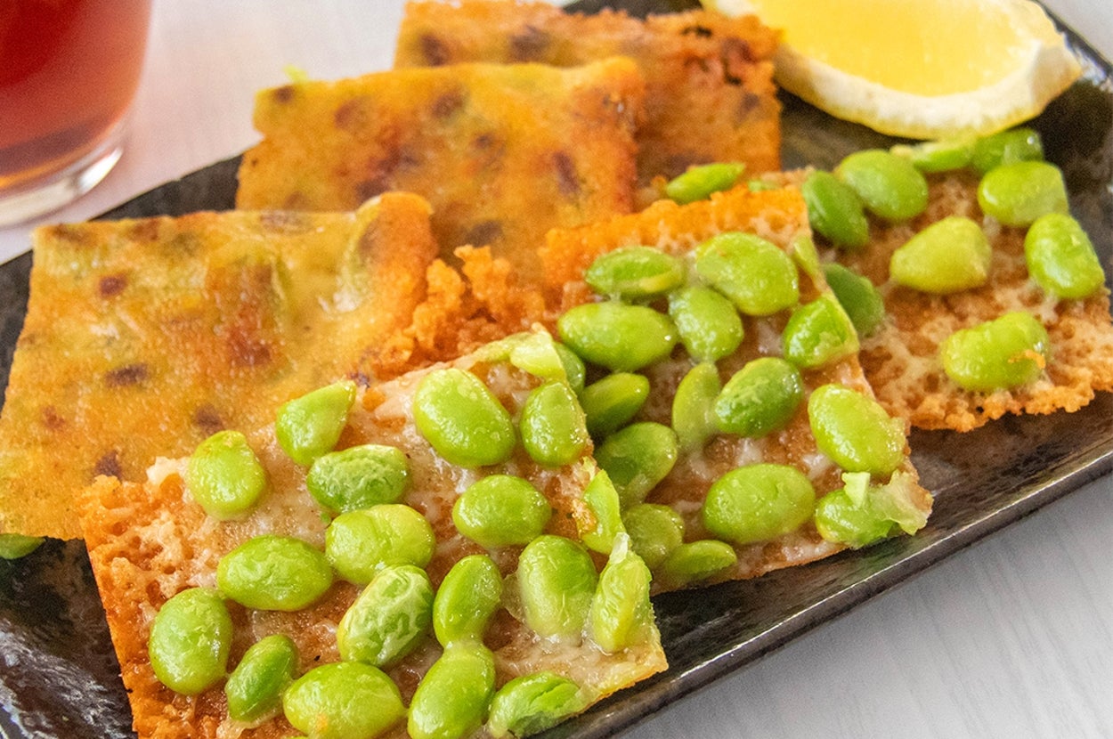 黒い皿に枝豆とチーズのカリカリしたおつまみとレモンが添えられている。横には飲み物がある。