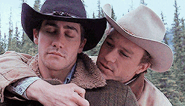 Two men in cowboy hats embrace outdoors, one resting his head on the other&#x27;s shoulder. A forest is visible in the background