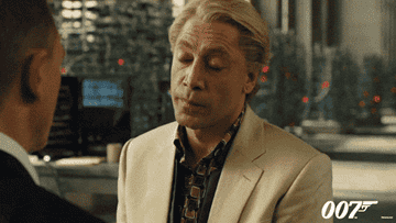 Two characters converse in a tense scene, one wearing a beige suit. The setting suggests a high-stakes situation from a spy film