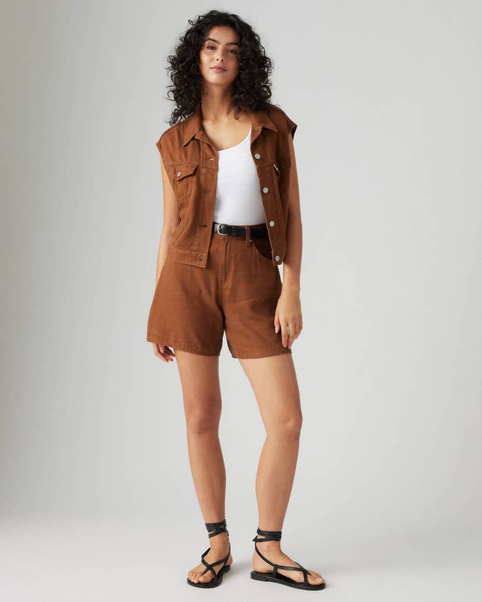 Woman with curly hair wearing a sleeveless brown vest and matching shorts, white top, and black sandals. Casual and stylish summer outfit