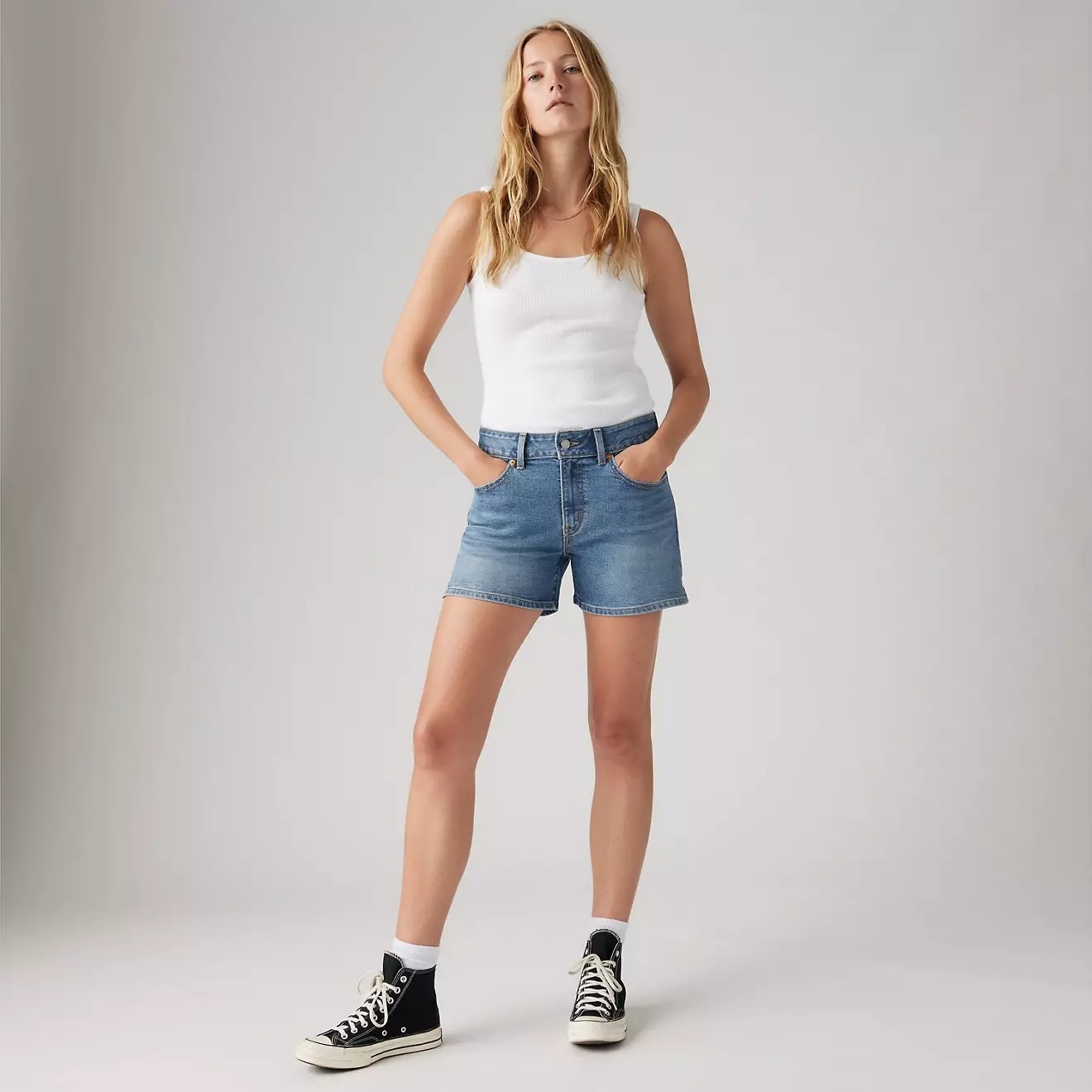 Person wearing a white tank top, denim shorts, and black high-top sneakers standing confidently with hands in pockets