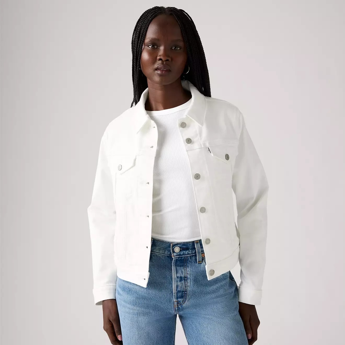 Person wearing a white denim jacket over a white shirt and blue jeans, posing confidently