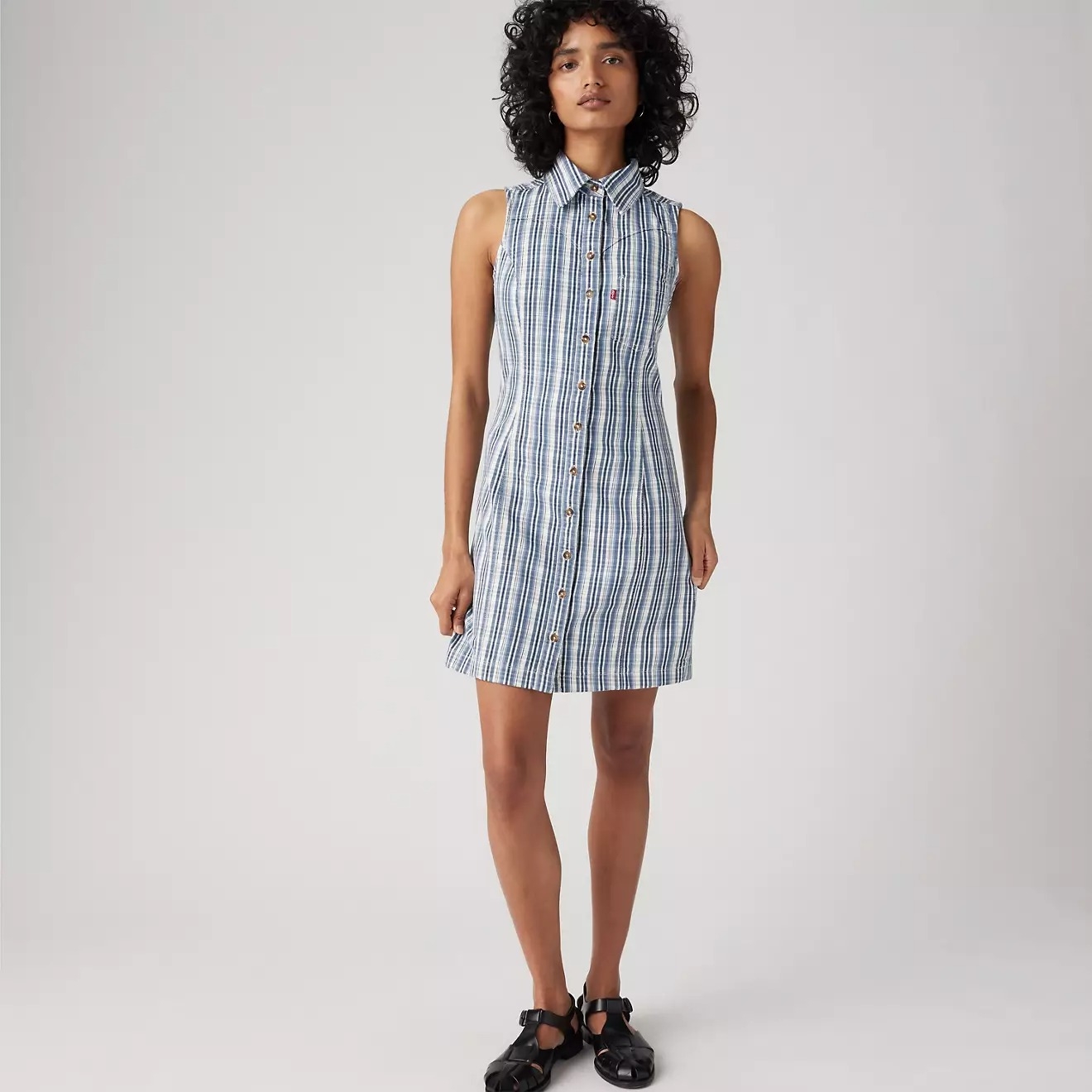 Woman wearing a sleeveless, button-up striped dress and black sandals, posing against a plain background
