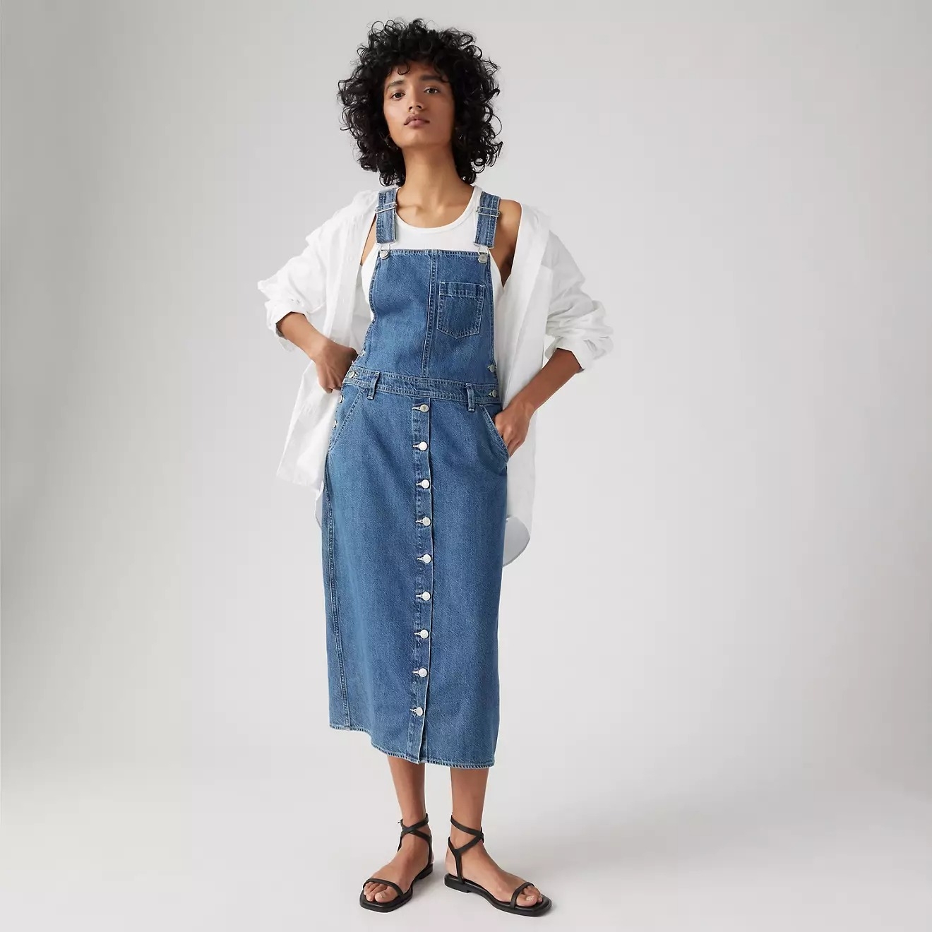 Person wearing a denim overall dress and sandals, with a white shirt underneath. Their hands rest in their pockets