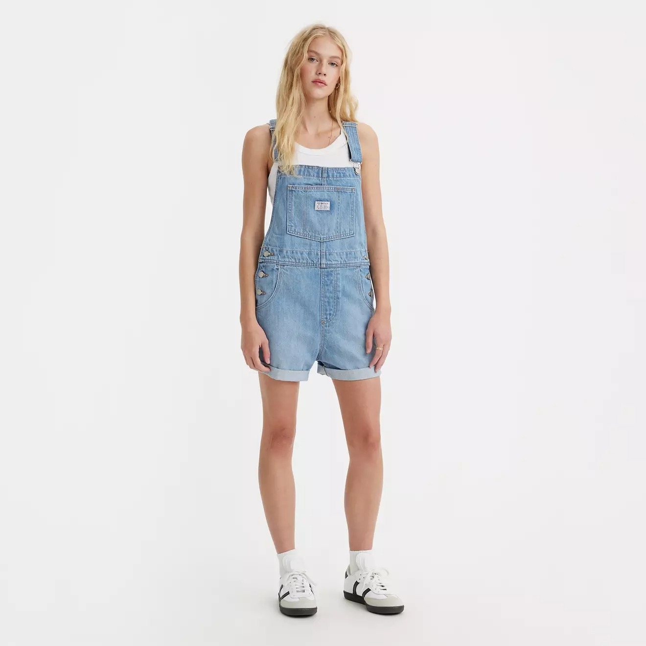 Person wearing denim overalls, white tank top, and sneakers, standing on a plain background