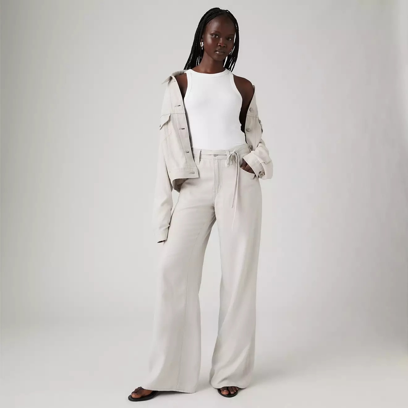 Model in a sleeveless white top, relaxed beige trousers, and matching jacket draped over shoulders, poses for a fashion shoot