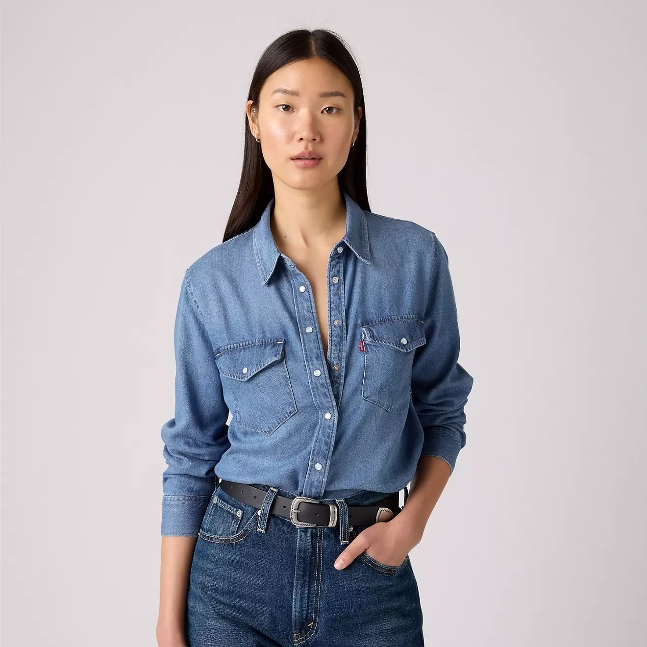 Person wearing a denim shirt with long sleeves, styled casually with faded jeans, standing with one hand in pocket