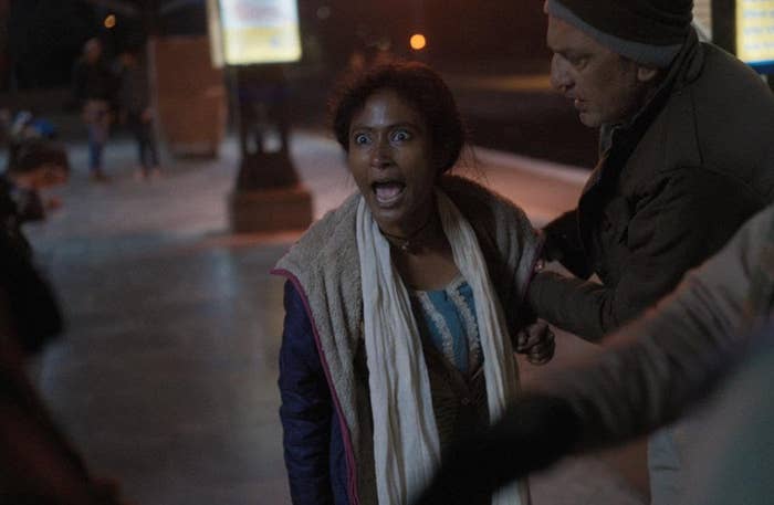 A woman standing on a platform appears shocked, turning towards the camera while a man beside her holds her arm