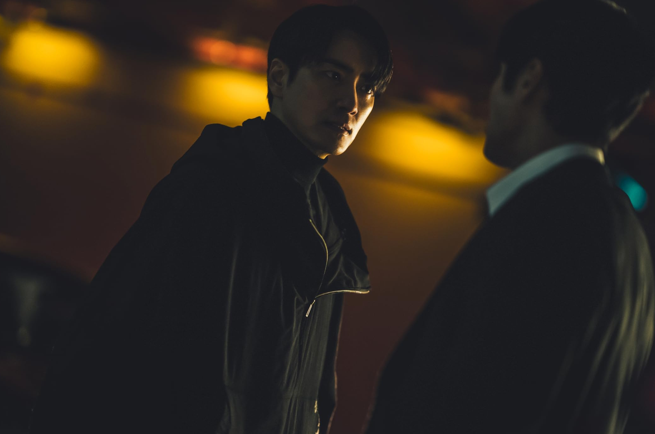 Two people in suits stand facing each other in a dimly lit room, creating a tense atmosphere. One person's face is visible, appearing serious