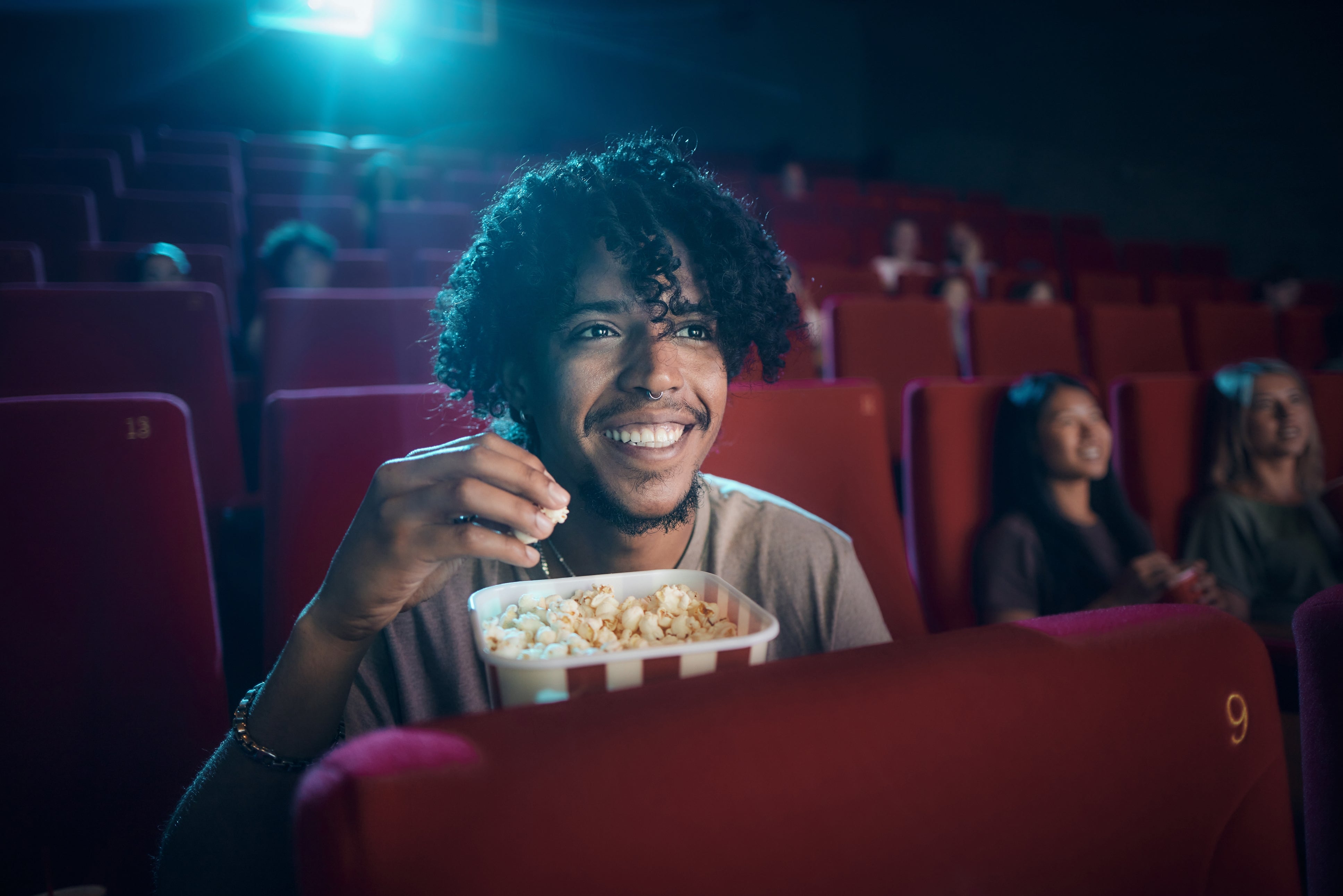 Person in a cinema smiling and holding a popcorn bucket, with others seated in the background