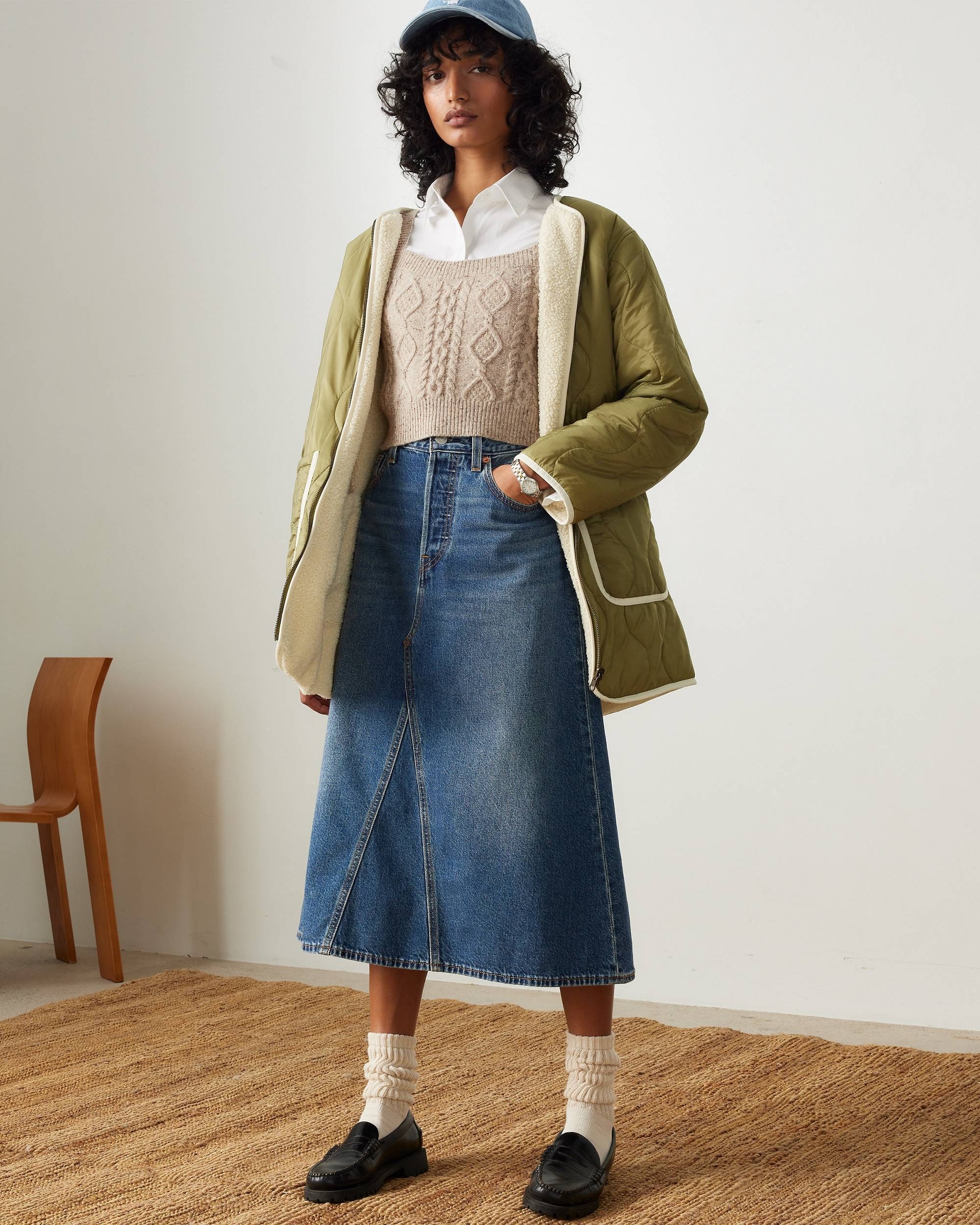 Person in casual autumn outfit: quilted coat, knit vest over white shirt, denim skirt, socks, loafers, and cap, standing on a textured rug