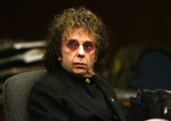 A person with curly hair and pink-tinted glasses is seated, wearing a dark suit, looking towards the camera