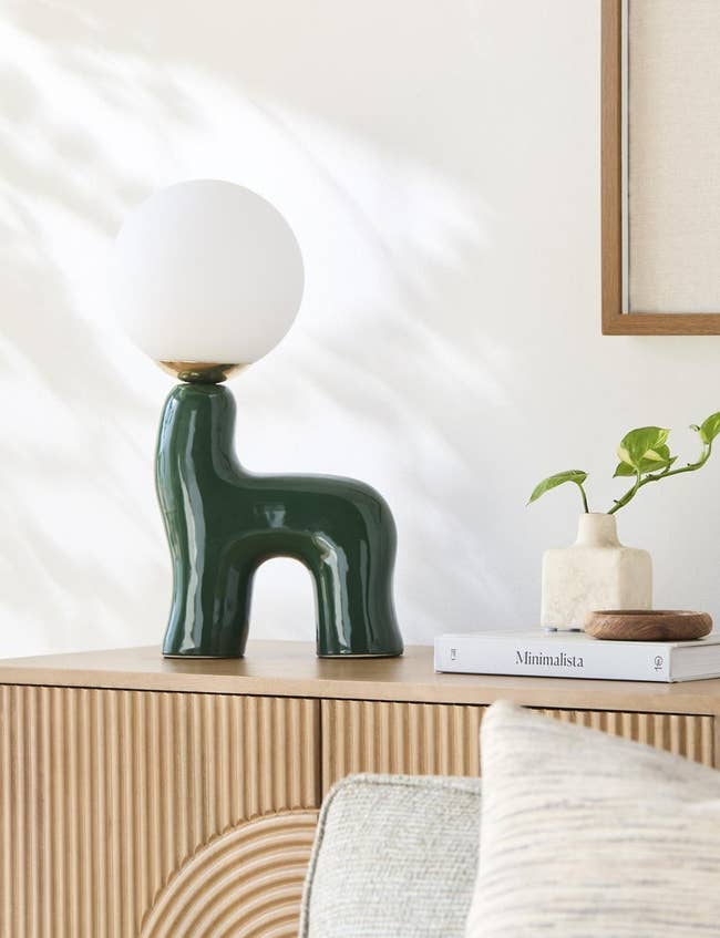 Modern array lamp with a achromatic globe connected a unique, greenish ceramic basal connected a woody furniture adjacent to a minimalist publication and a tiny plant