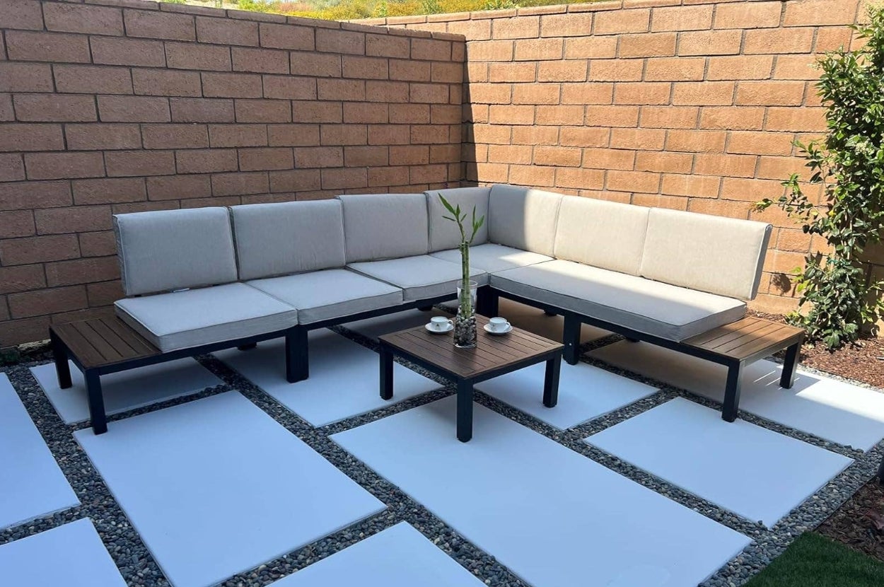 Outdoor patio with L-shaped beige sectional sofa, two wooden side tables, and a central coffee table on a modern stone tile floor