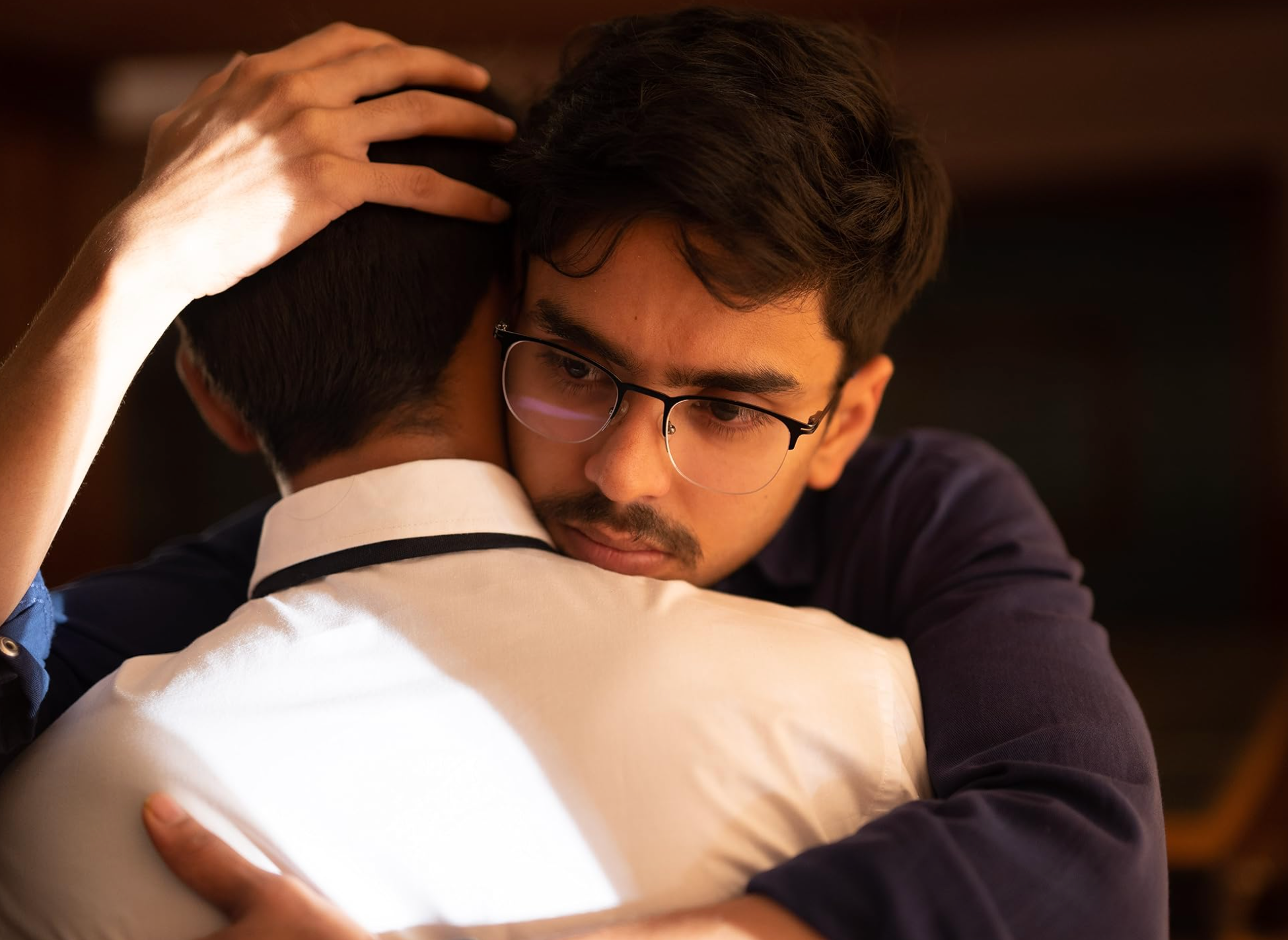 Two people share a sincere embrace. One wears glasses and a dark outfit; the other wears a light shirt. The mood is serious and comforting