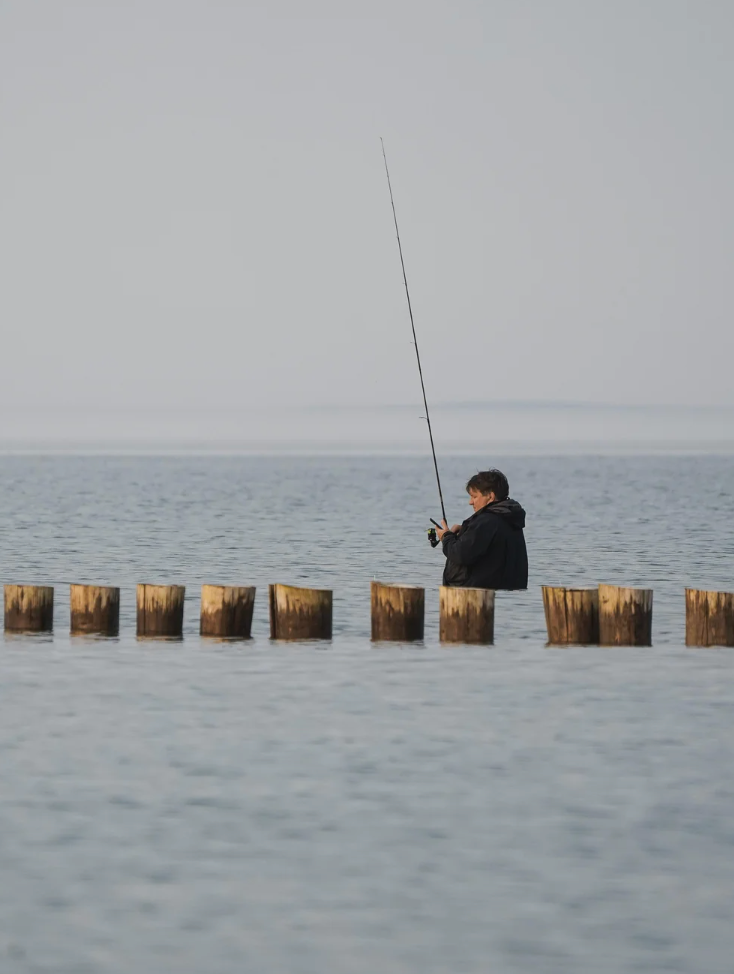 Person fishing from wooden posts in calm water, facing the open sea, under a cloudy sky