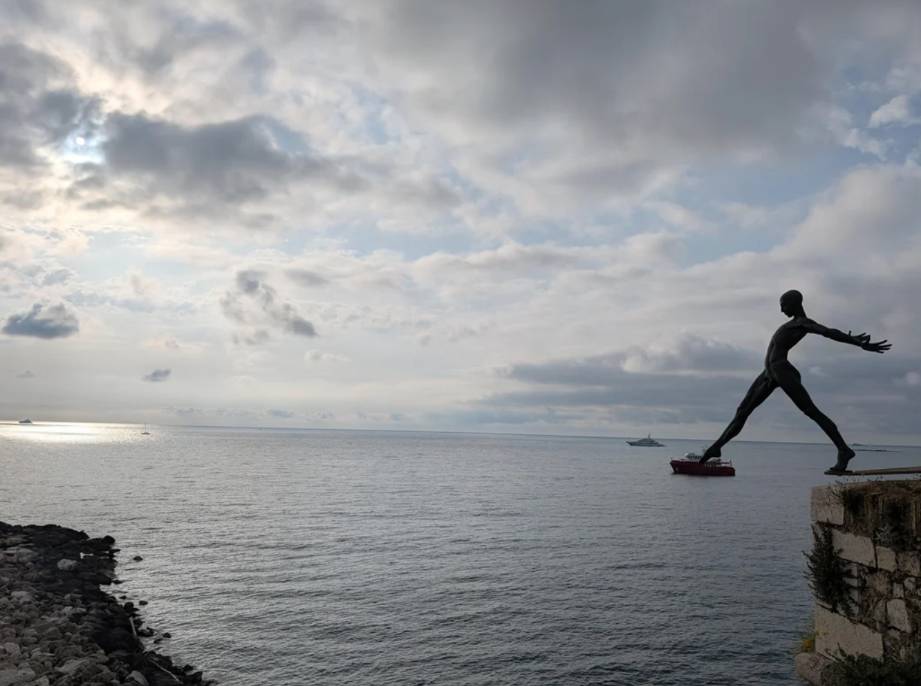 Sculpture of a figure poised on a ledge, overlooking a vast, calm sea under a cloudy sky