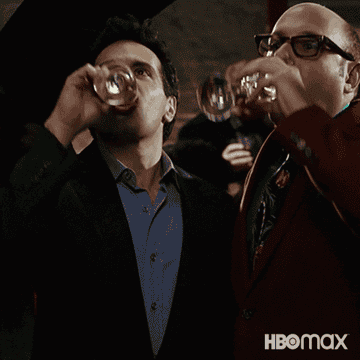 Two men in suits are confidently drinking from glasses at a lively gathering