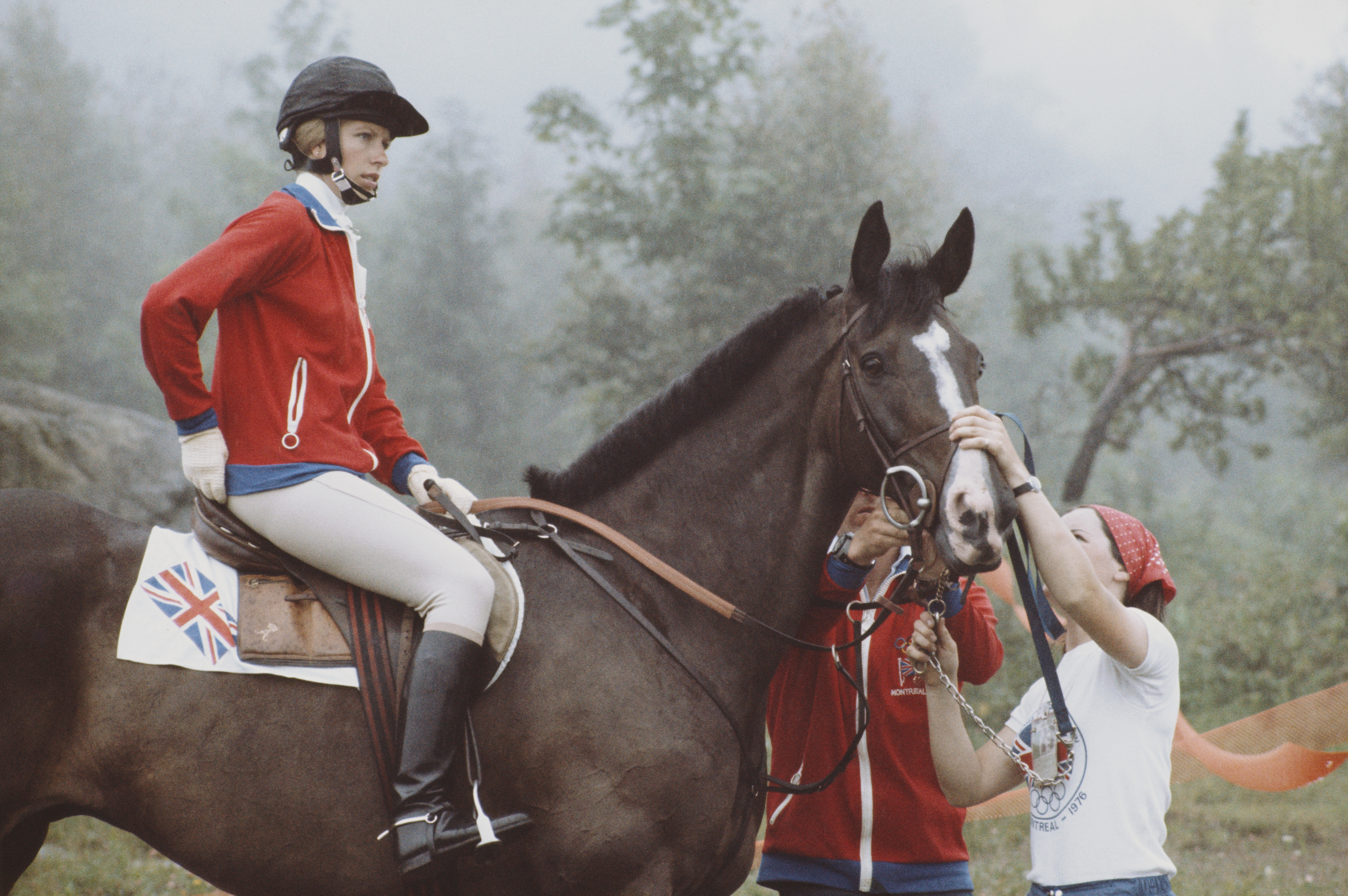 Person in equestrian gear sits on a horse, attended by two people. A British flag marks the horse's saddle area. Forested background visible