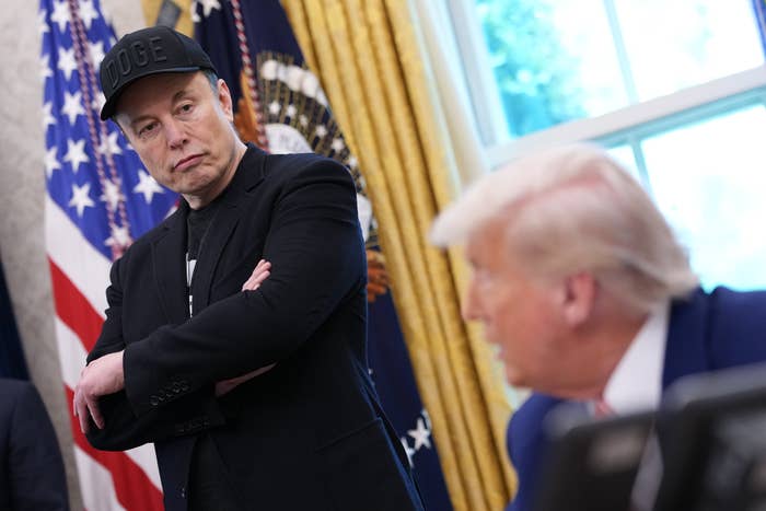 Two men in a formal setting, one with arms crossed wearing a cap, standing near flags; the other is seated, speaking