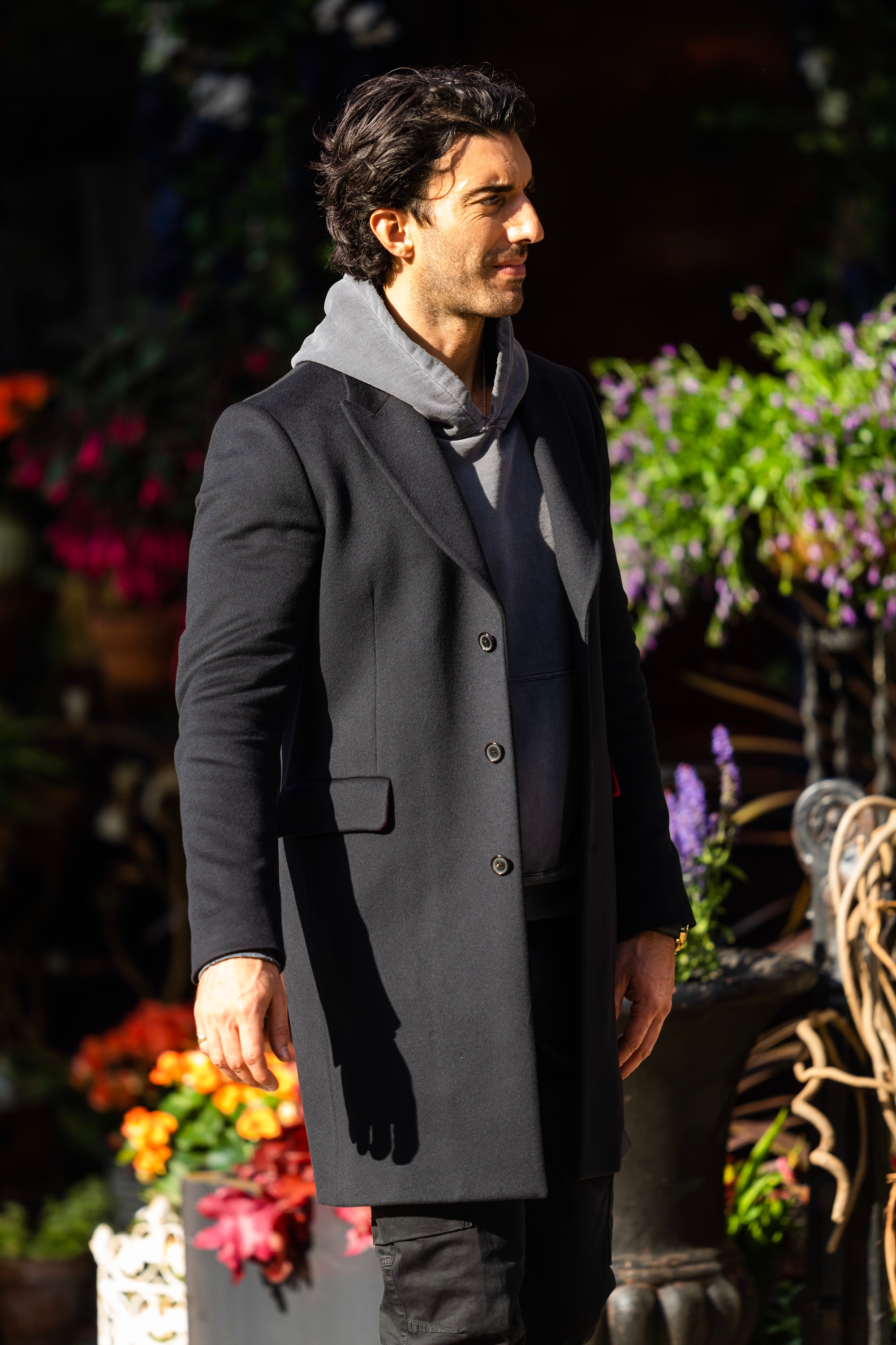 Justin Baldoni stands outdoors wearing a stylish coat over a hoodie, surrounded by vibrant plants and flowers
